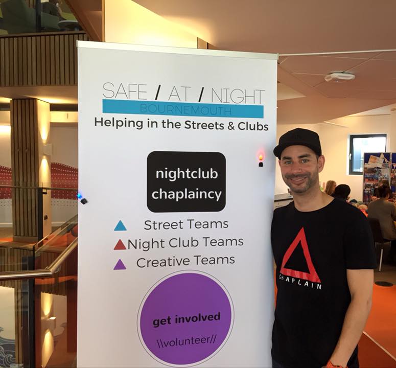 Kenny from the Nightclub Chaplaincy stands next to banner.